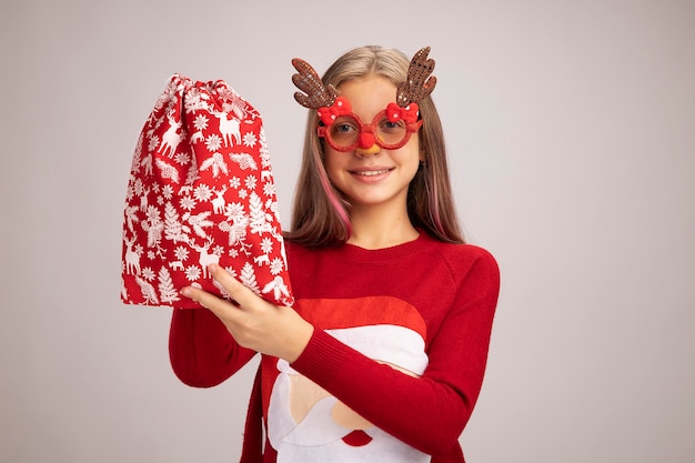 Fröhliches kleines Mädchen im Weihnachtspullover mit lustiger Partybrille, die eine rote Weihnachtstasche mit Geschenken hält und die Kamera fröhlich lächelt, die über weißem Hintergrund steht