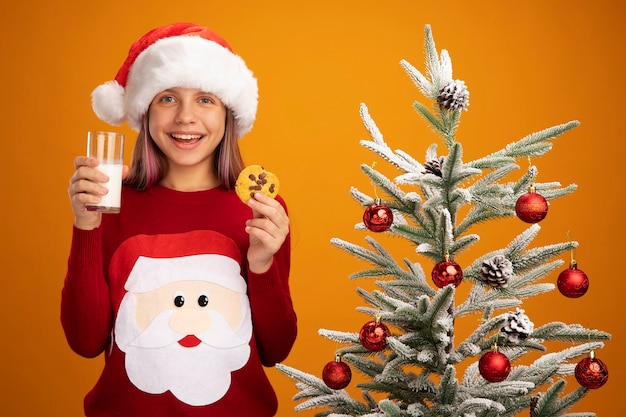 Fröhliches kleines Mädchen in Weihnachtspullover und Weihnachtsmütze mit einem Glas Milch und Keksen mit Blick auf die Kamera, die fröhlich neben einem Weihnachtsbaum auf orangefarbenem Hintergrund steht