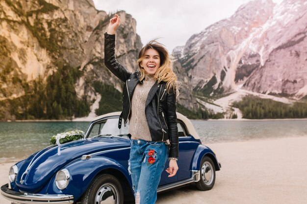 Fröhliches lachendes Mädchen, das Jeans trägt, die mit Stickerei verziert sind, die nahe dem Auto auf Berg aufwirft