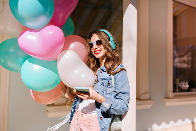 Fröhliches lächelndes Mädchen in der stilvollen Sonnenbrille, die zum Ereignis geht und Lieblingsmusik in den Kopfhörern hört. Entzückende junge Frau, die Retro-Jeansjacke trägt, die bunte Luftballons zur Geburtstagsfeier trägt.