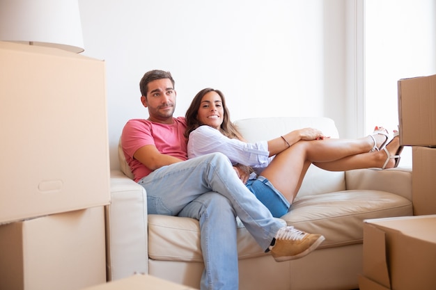 Fröhliches lateinisches junges Paar, das auf Couch unter Papppaketen in der neuen Wohnung sitzt,