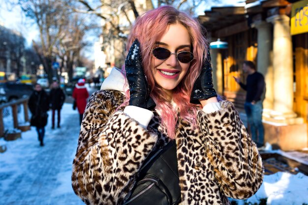 Fröhliches Lifestyle-Porträt der hübschen Frau mit ungewöhnlichen rosa Haaren im Freien, das eine trendige Leopardenfelljacke, eine Sonnenbrille im Vintage-Stil der 90er Jahre und eine Gürteltasche trägt, Grunge Street Wear, Wither Snow City.