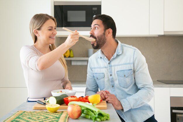 Fröhliches lustiges junges Paar, das Abendessen zusammen kocht und frisches Gemüse in der Küche aufschneidet. Frau, die ihrem Freund ein Stück Essen auf einem großen Löffel zum Schmecken gibt. Familienkochkonzept