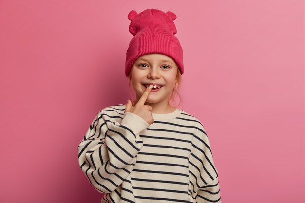 Fröhliches lustiges kleines Mädchen zeigt auf ihre Zähne, kümmert sich um Mundhygiene, trägt modische Kleidung, hat gesunde Haut, rühmt sich von erwachsenen Zähnen an Freunde auf dem Spielplatz, isoliert auf rosa Pastellwand