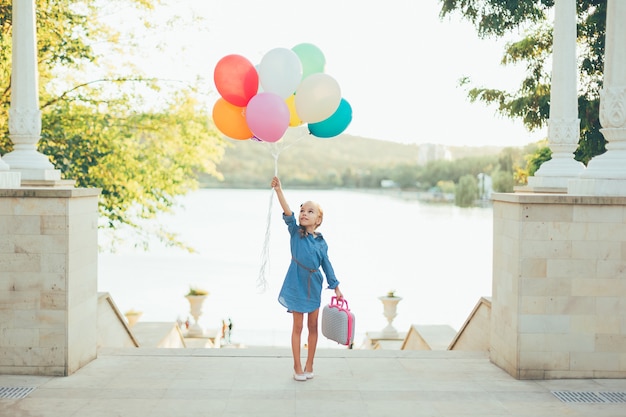 Fröhliches Mädchen, das bunte Luftballons und kindlichen Koffer hält