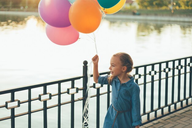 Fröhliches Mädchen, das bunte Luftballons und kindlichen Koffer hält