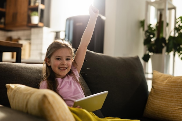 Fröhliches Mädchen, das mit erhobenem Arm feiert, während es im Wohnzimmer Spiele auf dem Touchpad spielt