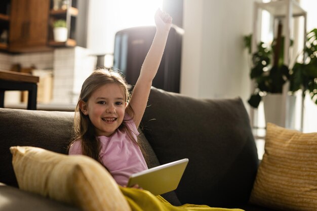 Fröhliches Mädchen, das mit erhobenem Arm feiert, während es im Wohnzimmer Spiele auf dem Touchpad spielt