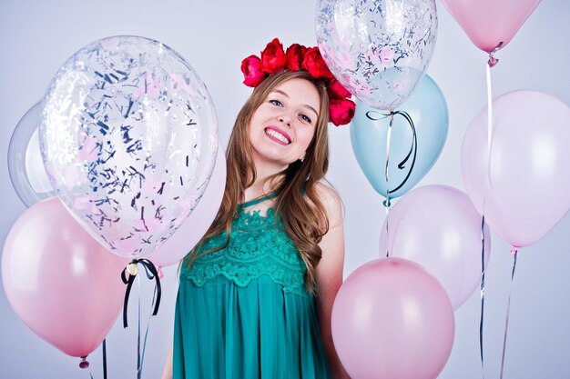 Fröhliches Mädchen in grünem türkisfarbenem Kleid und Kranz mit farbigen Luftballons isoliert auf Weiß Geburtstagsthema feiern