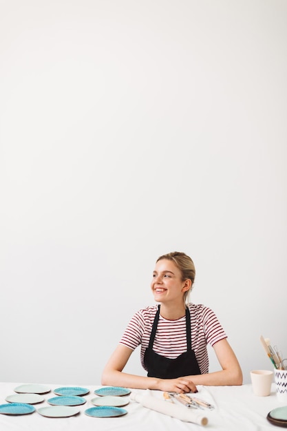 Fröhliches Mädchen in schwarzer Schürze und gestreiftem T-Shirt, das mit handgefertigten Tellern am Tisch sitzt und glücklich in das Töpferstudio blickt