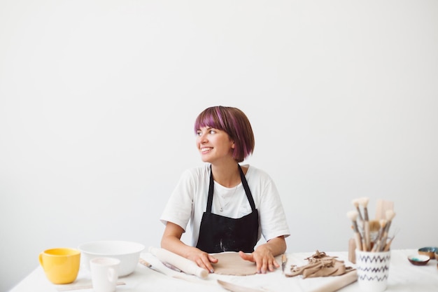 Fröhliches Mädchen mit bunten Haaren in schwarzer Schürze und weißem T-Shirt, das mit Ton arbeitet und glücklich auf das moderne Töpferstudio blickt