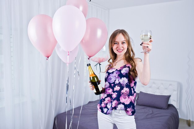 Fröhliches Mädchen mit farbigen Luftballons auf dem Bett im Zimmer mit Brille und Flasche Champagner Geburtstagsthema feiern