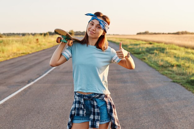 Fröhliches Mädchen mit stilvollem Haarband, das beim Gehen auf der Straße mit einem Longboard über den Schultern posiert, die Kamera mit glücklichen Emotionen anschaut und den Daumen zeigt.