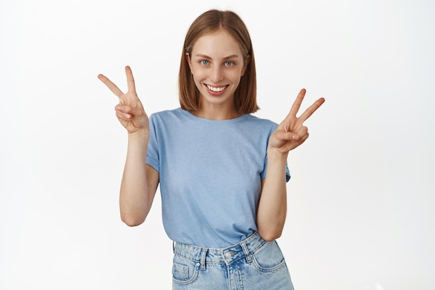 Fröhliches modernes blondes Mädchen, das V-Zeichen des Friedens zeigt und kawaii lächelt, Positivität, Spaß und Freude ausdrückt und im Sommer-T-Shirt auf weißem Hintergrund steht.