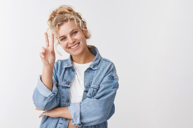 Fröhliches optimistisches, attraktives blondes europäisches Mädchen mit lockigem Brötchen-Haarschnitt zeigen Siegesfriedensgeste, die cool bleibt und Spaß hat, zu lächeln und Glück positive Einstellung zum Ausdruck zu bringen, weiße Wand