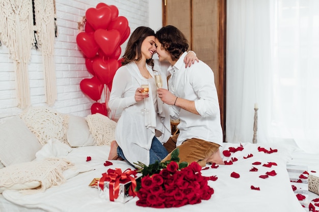 Fröhliches Paar sitzt auf dem Bett von Angesicht zu Angesicht mit Sektgläsern und feiert zusammen den Valentinstag