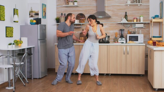 Fröhliches Paar tanzt und singt beim Frühstück in der Küche im Schlafanzug. Sorglose Frau und Ehemann lachen Spaß haben lustig das Leben genießen authentische verheiratete Menschen positive glückliche Beziehung