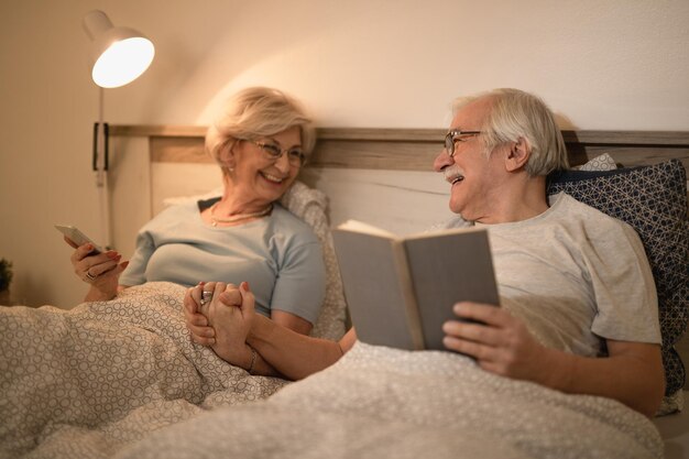 Fröhliches reifes Paar, das Händchen hält, während es sich im Bett ausruht und abends kommuniziert