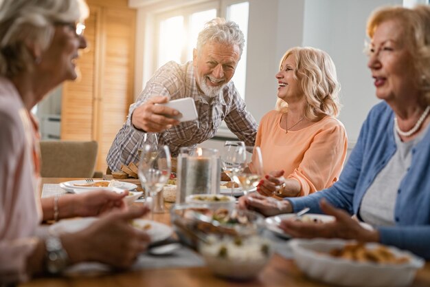 Fröhliches reifes Paar, das sich beim Mittagessen mit Freunden amüsiert und am Esstisch ein Selfie macht