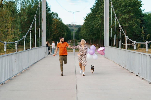 Fröhliches reizendes Paar, das glücklich auf Brücke mit ihrem lächelnden Hund und den rosa Luftballons läuft