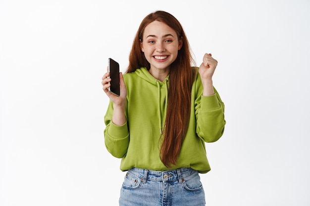 Fröhliches rothaariges Mädchen freut sich, hält Smartphone und feiert, gewinnt auf dem Handy und triumphiert, kauft mit Rabatt, steht auf Weiß