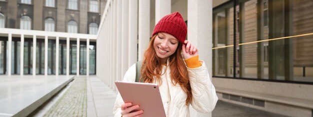 Fröhliches rothaariges Mädchen mit rotem Hut spaziert mit einem digitalen Tablet durch die Stadt und verbindet sich mit dem öffentlichen Internet-WLAN