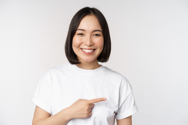 Fröhliches schönes asiatisches Mädchen, das Werbung auf leerem Kopierraum zeigt, mit dem Finger nach rechts zeigt und lächelt und Banner oder Firmenlogo auf weißem Hintergrund demonstriert