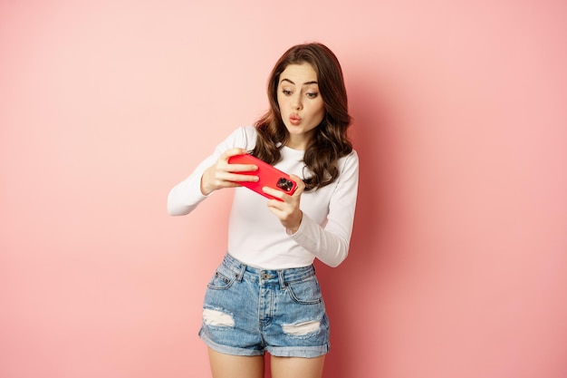 Fröhliches schönes Mädchen, das ein mobiles Videospiel spielt, das Smartphone horizontal hält und auf dem Handy mit aufgeregtem Gesicht, rosafarbenem Hintergrund zuschaut