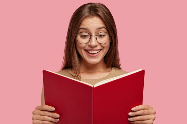 Fröhliches Schulmädchen hält Lehrbuch, liest interessantes Buch
