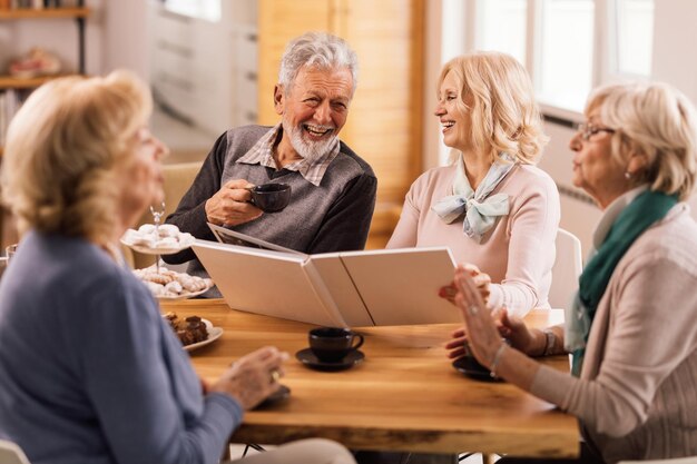 Fröhliches Seniorenpaar, das sich beim Betrachten des Fotoalbums mit seinen Freunden während der Kaffeezeit zu Hause amüsiert