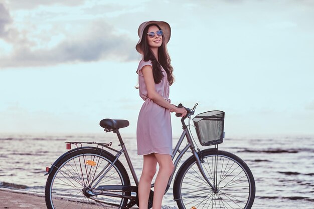 Fröhliches sinnliches Mädchen mit Sonnenbrille und Hut im Kleid geht mit ihrem Fahrrad am Strand vor erstaunlichem Küstenhintergrund spazieren.