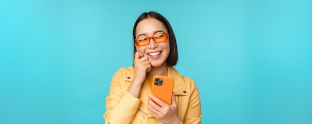 Fröhliches stilvolles asiatisches Mädchen mit Smartphone und lachendem Lächeln in die Kamera, die über blauem Hintergrund steht