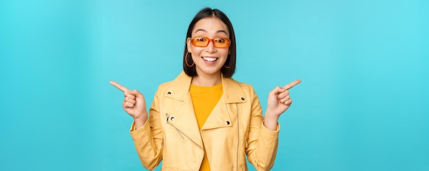 Fröhliches stilvolles chinesisches Mädchen mit Sonnenbrille zeigt mit den Fingern seitlich und lädt zum Auschecken ein