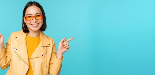 Fröhliches stilvolles chinesisches Mädchen mit Sonnenbrille zeigt mit den Fingern seitlich und lädt zum Auschecken ein