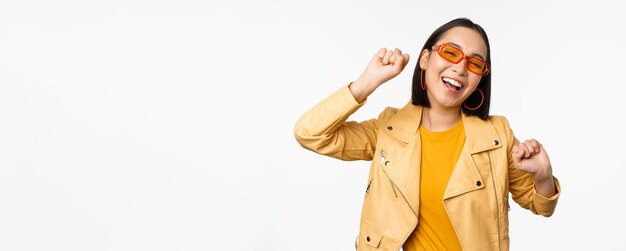 Fröhliches stilvolles koreanisches Mädchen mit Sonnenbrille, das sorglos über weißem Hintergrund tanzt und lacht und lächelt Kopierbereich