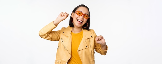 Fröhliches stilvolles koreanisches Mädchen mit Sonnenbrille, das sorglos über weißem Hintergrund tanzt und lacht und lächelt Kopierbereich