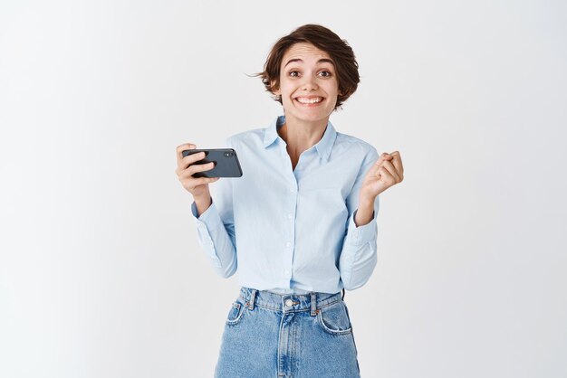 Fröhliches süßes Mädchen, das auf dem Handy gewinnt, Geld in der Smartphone-App verdient und lächelnd auf weißem Hintergrund steht