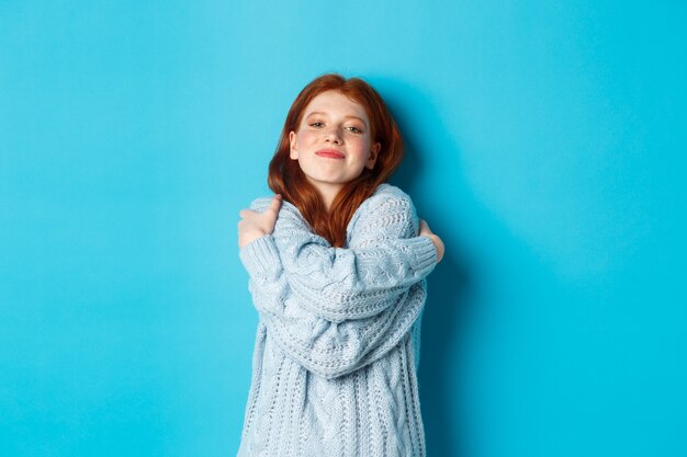 Fröhliches süßes rothaariges Mädchen, das sich selbst umarmt, einen bequemen und warmen Pullover trägt, in die Kamera lächelt und auf blauem Hintergrund steht.