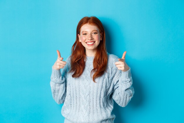 Fröhliches Teenager-Mädchen mit roten Haaren, das mit den Fingern auf die Kamera zeigt und Sie lächelt, gratuliert oder lobt und auf blauem Hintergrund steht