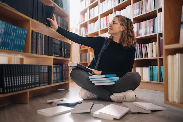 Fröhliches Teenager-Mädchen oder Studentin, die Bücher aus dem Regal in der Bibliothek nimmt - Menschen, Wissen, Bildung und Schulkonzept
