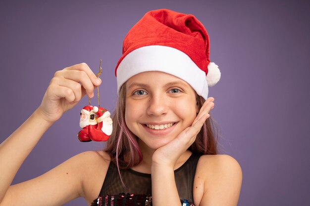 Fröhliches und zufriedenes kleines Mädchen in Glitzer-Partykleid und Weihnachtsmütze, das Weihnachtsspielzeug hält und in die Kamera schaut, die fröhlich über lila Hintergrund lächelt