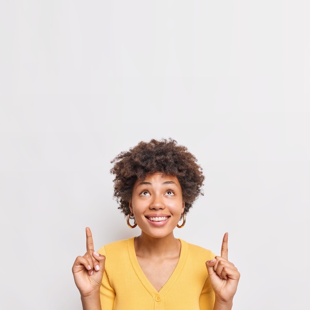 Fröhliches weibliches model mit lockigem haar zeigt nach oben in der werbung lächelt breit zeigt sonderangebot oder artikel mit rabatt trägt gelbe pullover steht gegen weiße wand