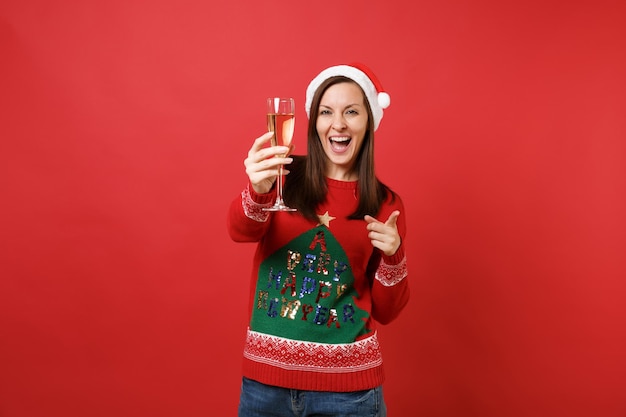 Fröhliches weihnachtsmädchen in weihnachtsmütze, das mit dem zeigefinger auf die kamera zeigt und ein glas champagner auf rotem hintergrund hält. frohes neues jahr 2019 feier urlaub party konzept. kopieren sie platz.