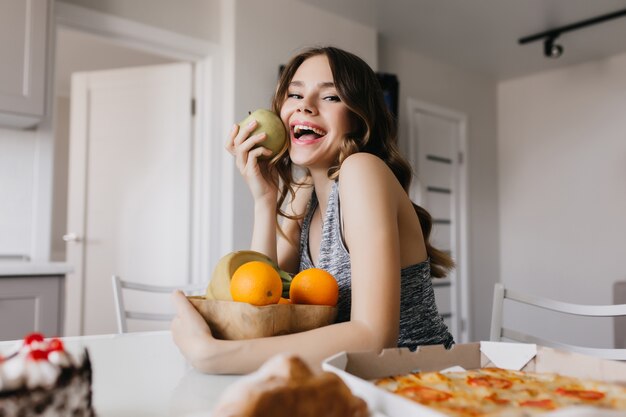 Fröhliches weißes Mädchen, das leckeren Apfel und Orange isst. Romantisches weibliches Modell, das Diät mit gesundem Essen genießt.