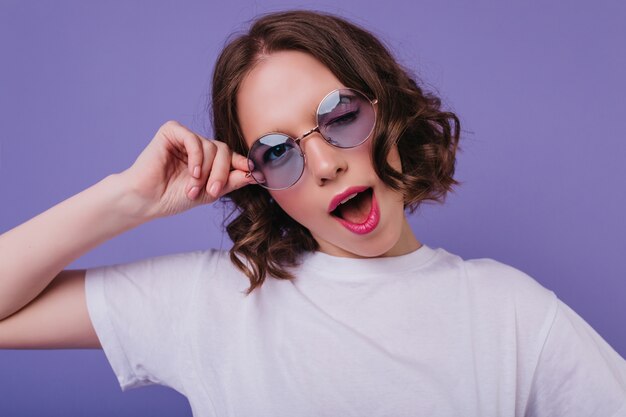 Fröhliches weißes Mädchen in der Sonnenbrille, die spielerisch auf hellvioletter Wand aufwirft. Innenfoto der sinnlichen lockigen jungen Frau.