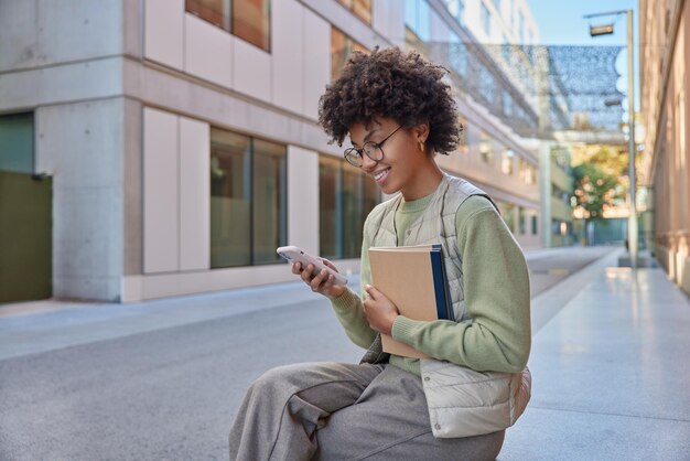 Froh, dass der Student lockiges Haar hat, hält ein Notebook, das Mobiltelefon zum Surfen im Internet verwendet, liest Ideen für ein Projekt, das lässig gekleidet im Freien posiert