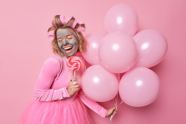 Froh, dass junge Frau Ton-Schönheitsmaske zur Hautbehandlung aufträgt, Karamellbonbons auf einem Stock hält und ein Bündel aufgeblasener Heliumballons trägt, festliches Kleid macht, Frisur isoliert über rosa Hintergrund