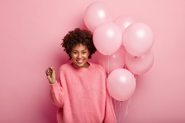 froh, dass lockiges weibliches Model die Faust ballt, einen übergroßen Pullover trägt, Luftballons hält, glücklich, bei der Geburtstagsfeier anwesend zu sein, einen rosa Pullover in einem Ton mit Wand trägt.