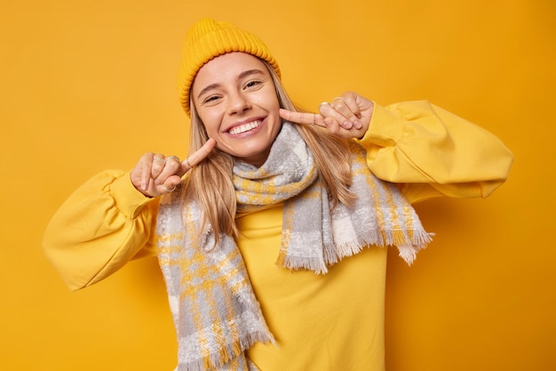 Kostenloses Foto froh, junge frau zeigt auf zahniges lächeln, hat weiße, gleichmäßige zähne, trägt ein hut-sweatshirt und einen schal um den hals, fühlt sich isoliert über gelbem studiohintergrund sehr glücklich. konzept der menschlichen gesichtsausdrücke