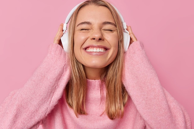 Froh Millennial Mädchen schließt die Augen und grinst in die Kamera hört Lieblingsmusik über Stereokopfhörer trägt bequemen weichen Pullover isoliert über rosa Hintergrund People Lifestyle und Hobby Konzept